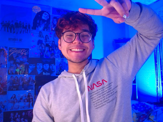 EddTerence, with curly brown hair and glasses, smiling with eyes closed, wearing a grey NASA hoodie, making a peace sign with his right hand, has a cute face and a small beard.