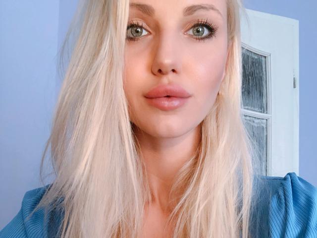 Lilith91-hot has blonde hair, green eyes, and pink lips with a slight pout. She's wearing a blue shirt, and her tits are partially visible. Her face is centered.