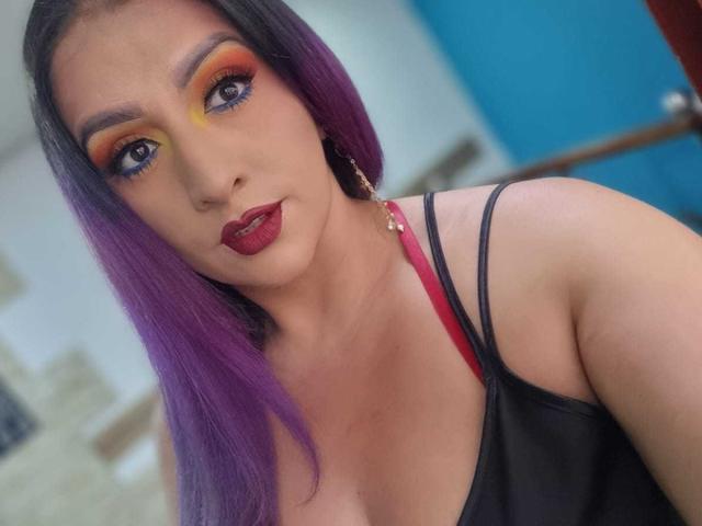 AndreaSharick with purple hair, red lipstick, orange-yellow eyeshadow, and black strappy top, showing off her tits, wearing gold earrings, and looking at the camera.