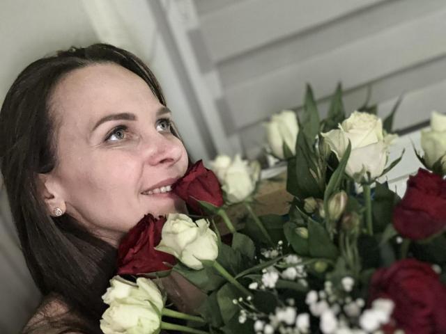 DesireMari with dark brown hair, holding red and white roses, smiling with a red rose in her mouth, looking to the side, wearing small diamond earrings.