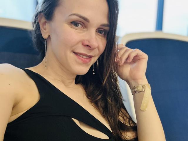 DesireMari with wet brown hair, black cut-out dress, and bracelet, smiling at the camera, showing a bandage on her wrist, wearing dangling earrings, looking cute.