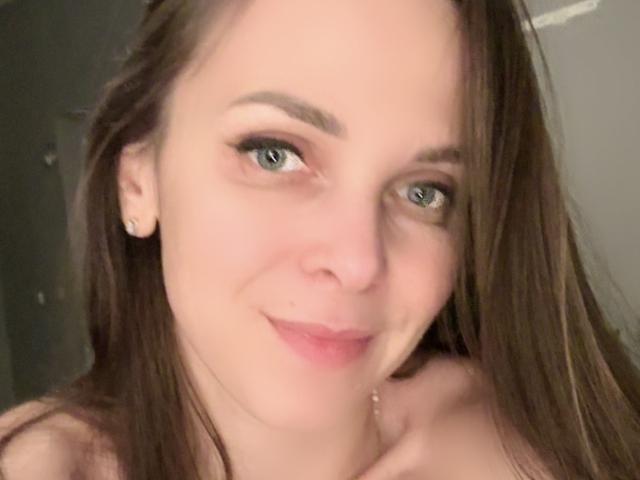 DesireMari's face, showing light skin, green eyes, brown hair, and a subtle smile, with small earrings, looking slightly to the right. Her tits are not visible.