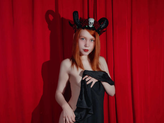 CheryCake, avec une peau pâle, des cheveux roux, une tête ornée de cornes noires, un crâne, et un tissu noir recouvrant ses seins, debout devant un rideau rouge, regardant la caméra, avec une ombre sur le rideau..