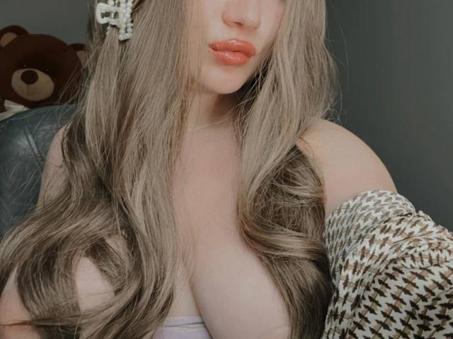 IrenRori with long, wavy, blonde hair, orange lipstick, and a brown and white houndstooth shirt, showing off her big, pale, bare tits. A brown teddy bear in the background.