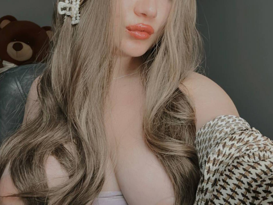 IrenRori with long, wavy, blonde hair, orange lipstick, and a brown and white houndstooth shirt, showing off her big, pale, bare tits. A brown teddy bear in the background.
