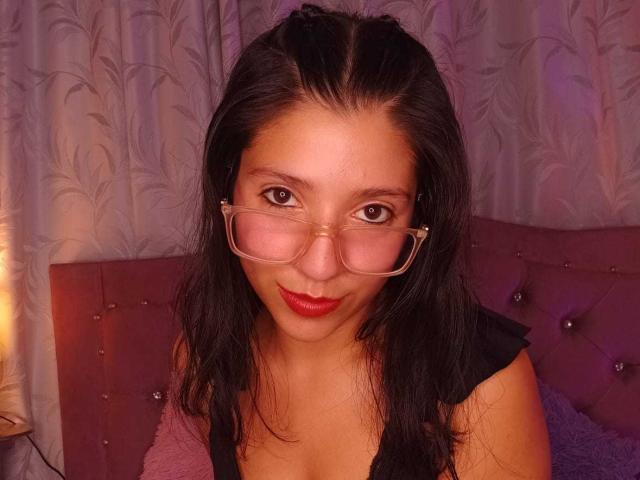 GagPrinceess con capelli scuri, occhiali e rossetto rosso, indossa una maglietta nera, seduta su una testiera viola imbottita, sorridendo alla fotocamera. Il seno non è visibile..