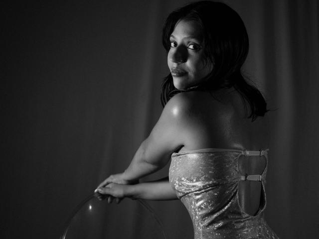 GagPrinceess with dark hair, looking over shoulder, wearing a shiny strapless dress, holding a clear, curved object, black-and-white photo, showing her back and slightly turned face.