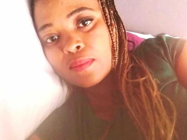 Ellerie with brown braids, pink lips, and green eyes, lying on a white pillow, wearing a green shirt, looking straight at the camera with a slight pout.
