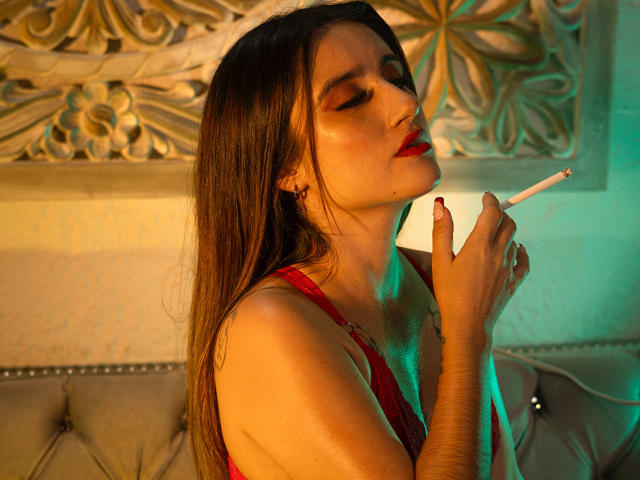 LindaSpark with long brown hair, red lipstick, and red lingerie, smoking a cigarette, eyes closed, holding it to her lips, green and yellow lighting, sitting on a tufted couch.