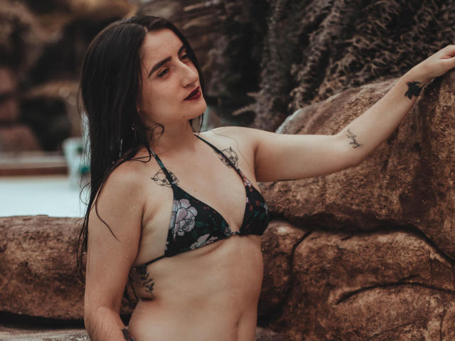 LindaSpark, con su largo cabello negro, vestida con un bikini floral, tiene tatuajes en el brazo y en el costado, apoyada en las rocas, mirando a la cámara con un lápiz labial rojo oscuro..