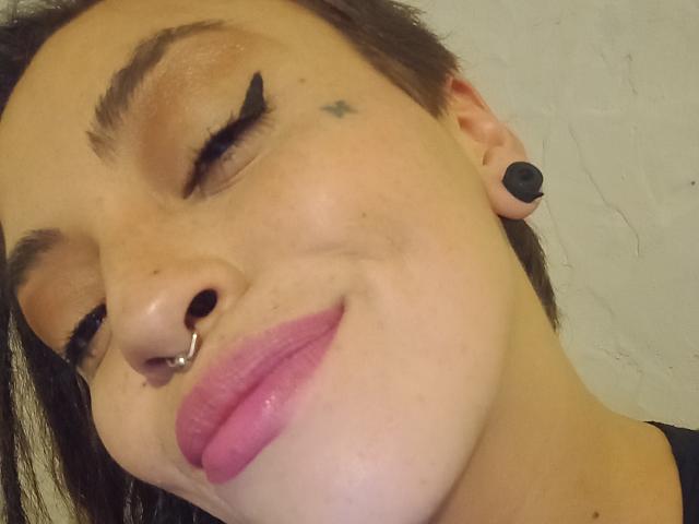 CattyStar has short brown hair, a nose ring, black ear plugs, and pink lipstick, with a small star tattoo near her right eye, smiling with eyes closed.