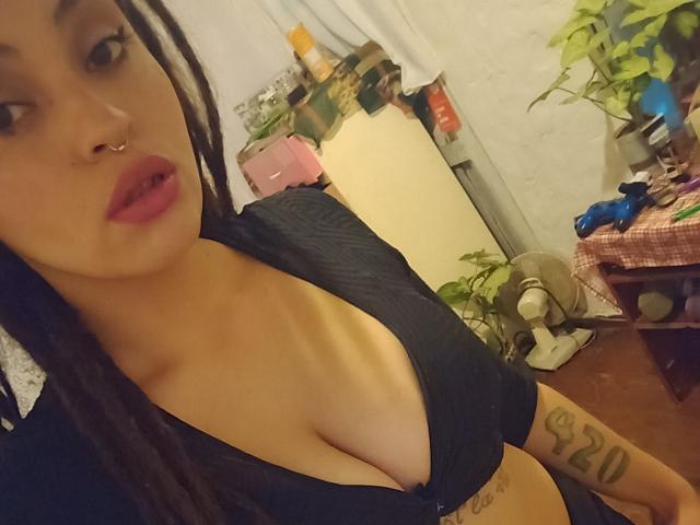 CattyStar with dark braided hair, nose ring, and pink lipstick, showing her big tits in a black top, tattoo on her right arm, in a dimly lit room.
