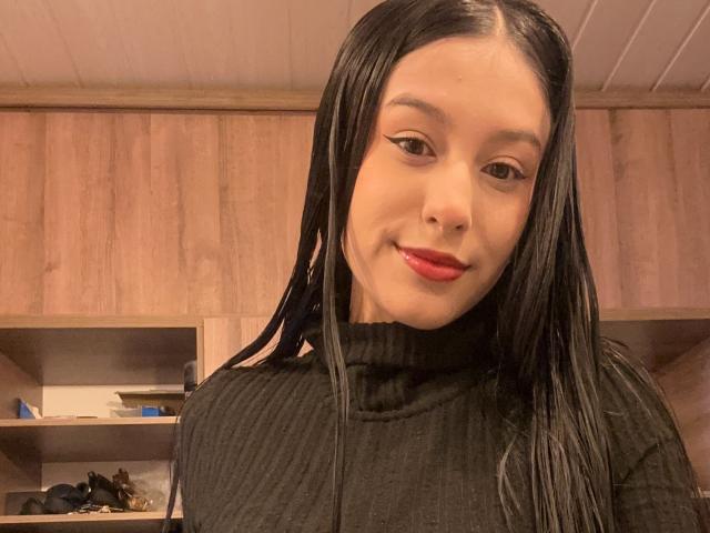 Soranaa with long black hair, red lipstick, and a black turtleneck, smiling slightly, in a wooden room, showing her smooth, light-skinned face and subtle makeup.