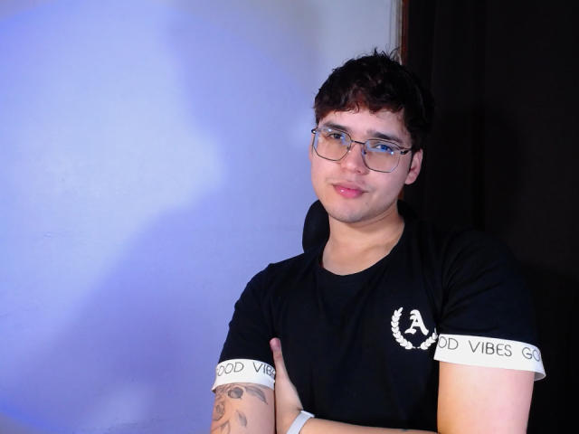 EddTerence with glasses, black tee, "Good Vibes" sleeves, tattooed arm, crossed arms, short dark hair, slight smile, white wristband, blue-purple light shadow.