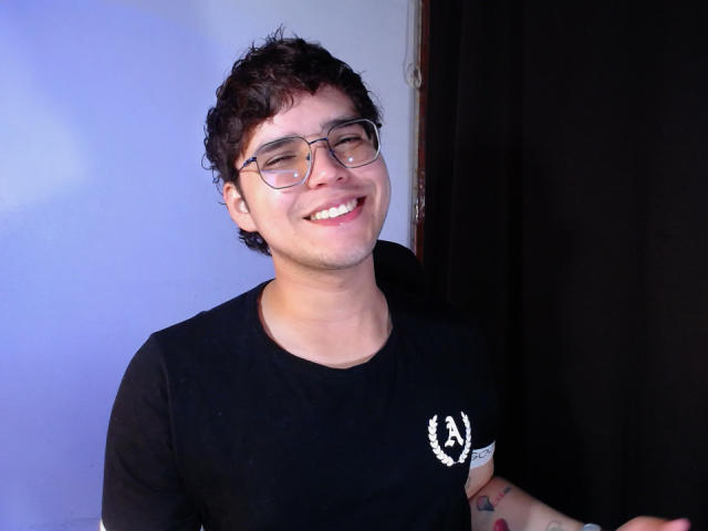 EddTerence with curly brown hair, glasses, smiling, wearing a black A-Team t-shirt, standing against a dark curtain, showing a tattoo on his right forearm.