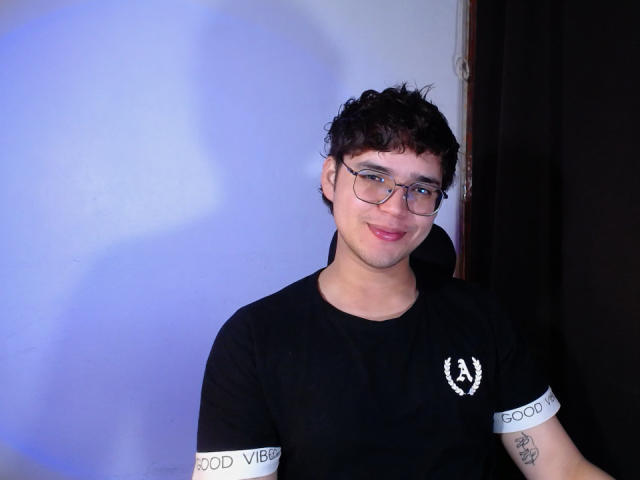 EddTerence, short curly dark hair, glasses, black t-shirt with white "GOOD VIBES" cuffs, tattoo on right forearm, slight smile, blue shadow on white wall.