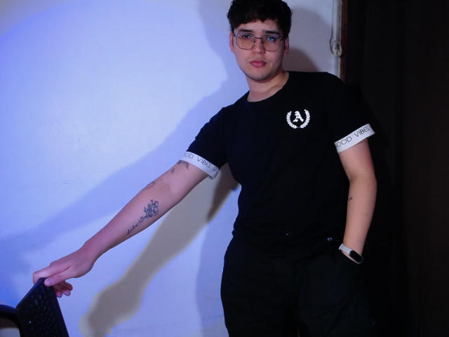EddTerence with short dark hair, glasses, black shirt with white sleeves, black pants, tattoo on left arm, wristwatch, leaning against wall, holding black bag.