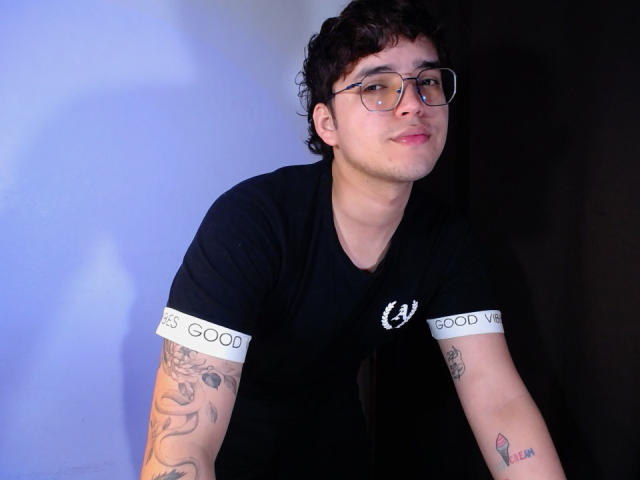 EddTerence with curly dark hair, glasses, and black Good Year t-shirt, leaning forward, showing tattoos on both arms, slight smile, white skin, looking at camera.