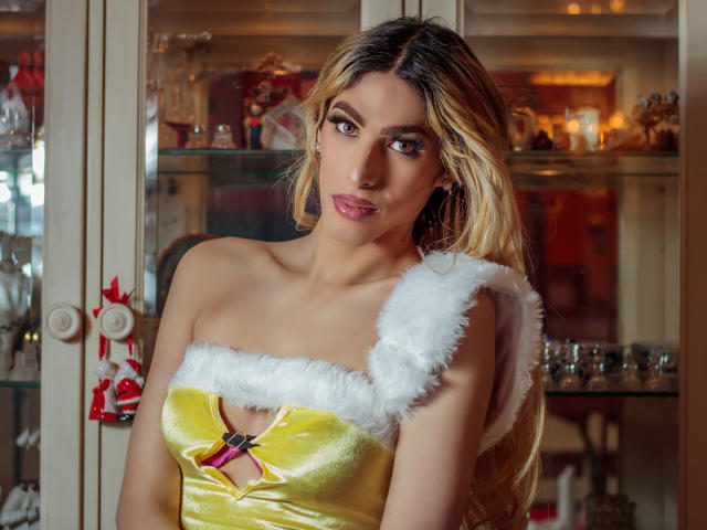 Putinna with blonde hair, wearing a yellow velvet dress with white fur trim, has medium tits, a cute face, and a sexy smile, standing in a cozy room.
