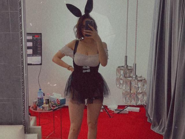 MonaLiss with bunny ears, white off-shoulder top, black skirt with fluffy butt, black bow, holding phone, standing in room, mirror selfie, showing her ass and tits.