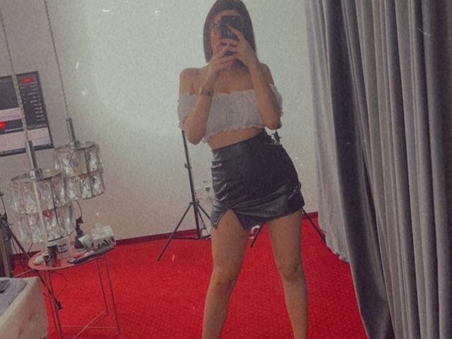 MonaLiss with off-shoulder white crop top and black leather skirt, holding phone, mirror selfie, standing on red carpet, gray curtains, and metal stands in background.