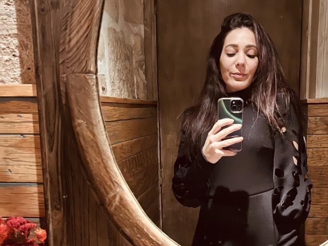 DiHotCouple with dark hair, eyes closed, wearing a black dress with cut-out sleeves, taking a selfie in a mirror, holding a green phone, wooden background, subtle smile.