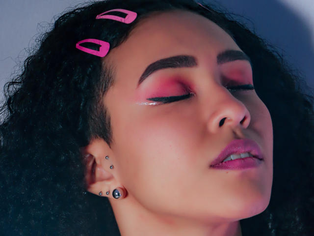 ZoeGruber has curly black hair with pink hairpins, pink eyeshadow, glossy pink lips, and multiple ear piercings, eyes closed, face slightly tilted up, looking relaxed.