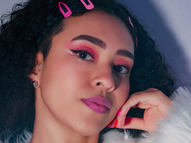 ZoeGruber with curly black hair, pink hair clips, and shiny makeup. She's got pink eyeshadow, glossy purple lips, and a piercing in her right ear. She's touching her face.