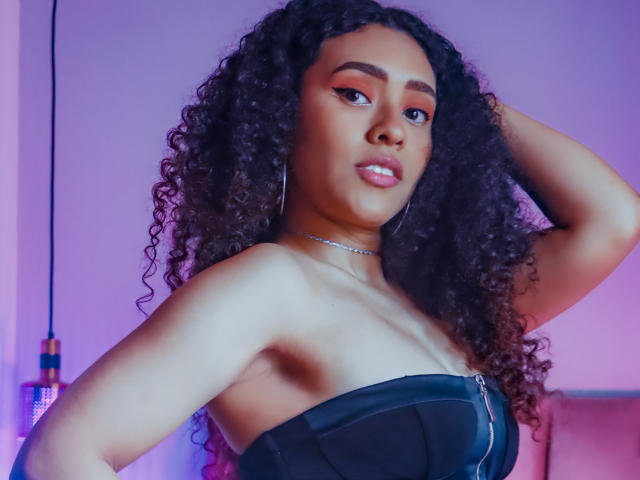 ZoeGruber with curly black hair, wearing a black strapless top, showing her bare shoulders and small tits, looking at the camera with pink eyeshadow and a silver necklace.
