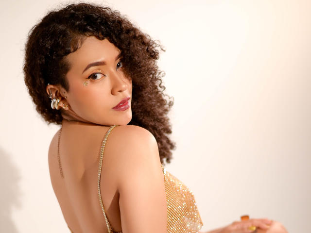 ZoeGruber with curly brown hair, tan skin, and gold chain dress, looking over her shoulder, showing her back and tiny gold stud on her cheek, with a seductive expression.
