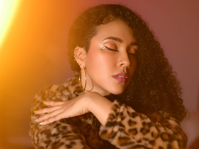ZoeGruber with curly dark hair, closed eyes, and gold eyeshadow, wearing a leopard print fur coat, large hoop earrings, and pink lipstick, has her hand on her chest.
