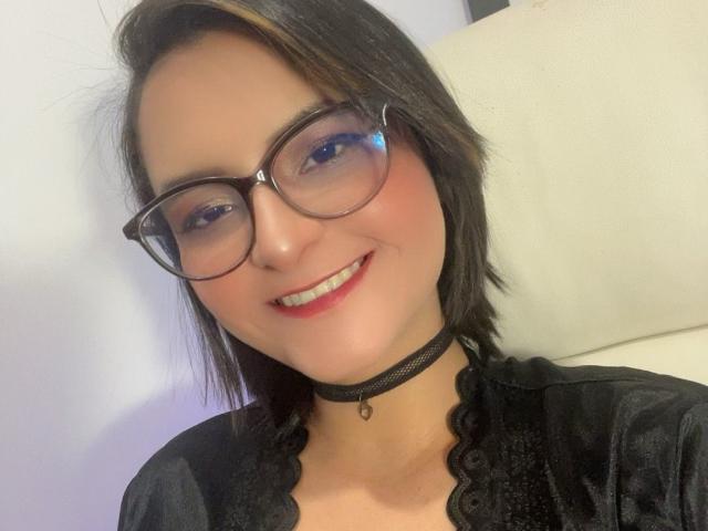 LeahKendl, con gafas, collarín negro y cabello castaño corto, sonriendo con lápiz labial rojo, vistiendo una blusa negra de encaje, mostrando un trasero lindo, pechos medianos y un rostro adorable..