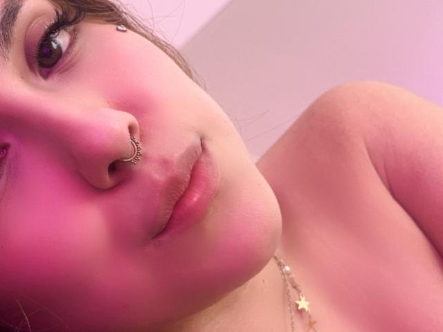 AmberlStha's pink-lit face with a nose ring, glossy lips, and a star necklace, showing her bare shoulder and a slight smile. Her skin is soft, and she's cute.