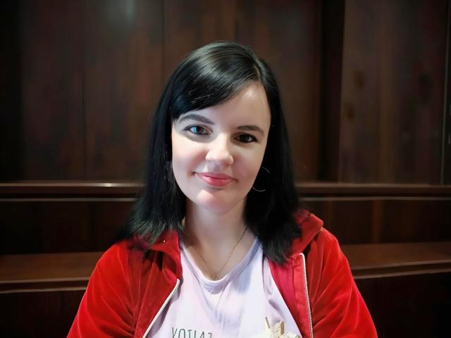 AlicexB with black hair, red hoodie, and white shirt, smiling slightly, hoop earrings, fair skin, brown eyes, medium build, sitting against dark wooden background.