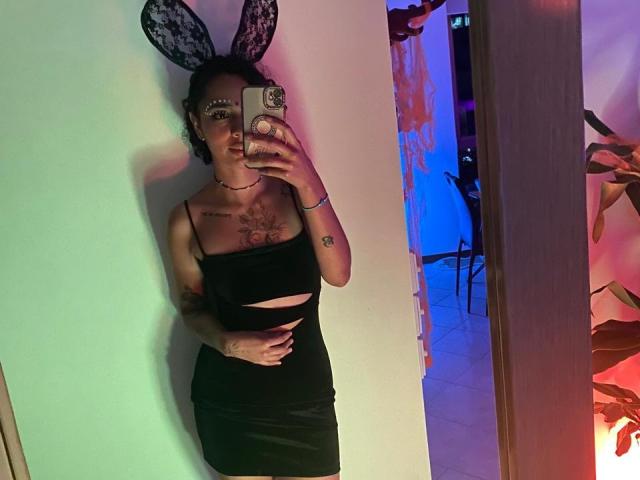 SilvanaXOwen, avec des oreilles de lapin en dentelle noire, des lunettes et une robe noire découpée, qui met en valeur ses seins et ses tatouages dans un selfie pris devant un miroir, tenant un téléphone avec un support pop..