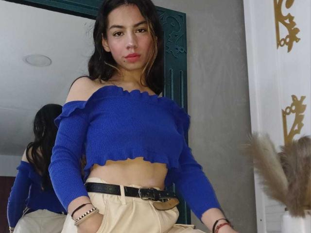 KatherinJohns with long dark hair, wearing a blue off-shoulder top, exposing her small tits, beige pants, black belt, and bracelet, standing in front of a mirror.