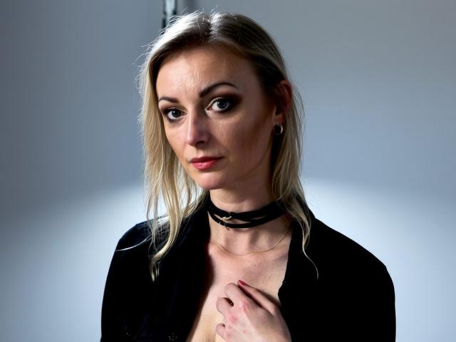 XLovelyVanessa has blonde hair, dark eyes, and a serious expression. She's wearing a black choker and blazer, with her hand on her chest, showing her tits slightly.