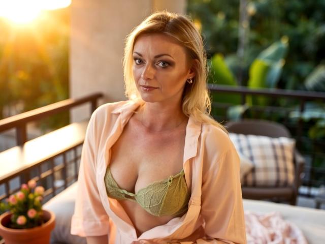 XLovelyVanessa with blonde hair, green lace bra, and pink shirt, showing her tits, sitting outside at sunset, looking slightly annoyed. Sunlight highlights her chest and face.
