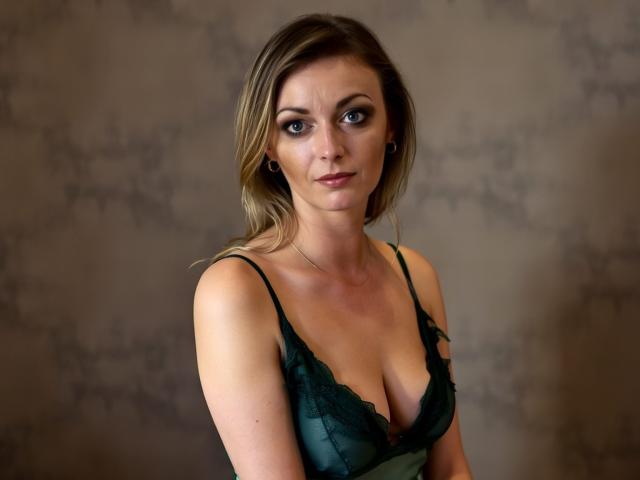 XLovelyVanessa with medium tits in a green lace bra, looking serious, has blonde hair, light skin, and small hoop earrings. She's centered in the image.