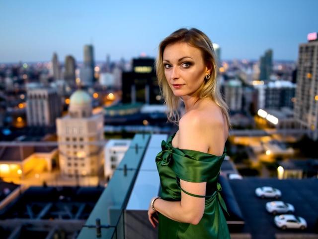 XLovelyVanessa with blonde hair, green off-shoulder dress, small tits, standing on rooftop, looking over city at dusk, blue sky, city lights, glass railing, city cars below.