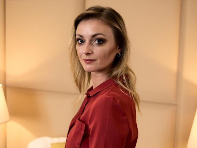 XLovelyVanessa with blonde hair, wearing a red shirt, looking over her shoulder, smiling subtly, brown eyes, dark makeup, small black earrings, in a softly lit room.