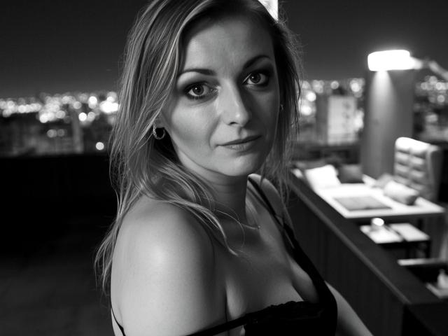 Black-and-white photo of XLovelyVanessa with shoulder-length blonde hair, small earrings, and a black lace top, looking directly at the camera with a slight smile. Her tits are visible.