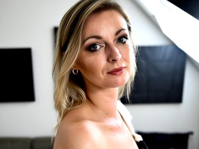 XLovelyVanessa has blonde hair, green eyes, and is looking over her shoulder with a slight smile. She's wearing small gold earrings and has a light skin tone.