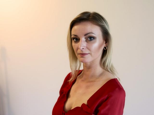 XLovelyVanessa with blonde hair, small tits, and a red dress with a deep cleavage, smiling slightly, wearing small hoop earrings, in a simple, well-lit photo.