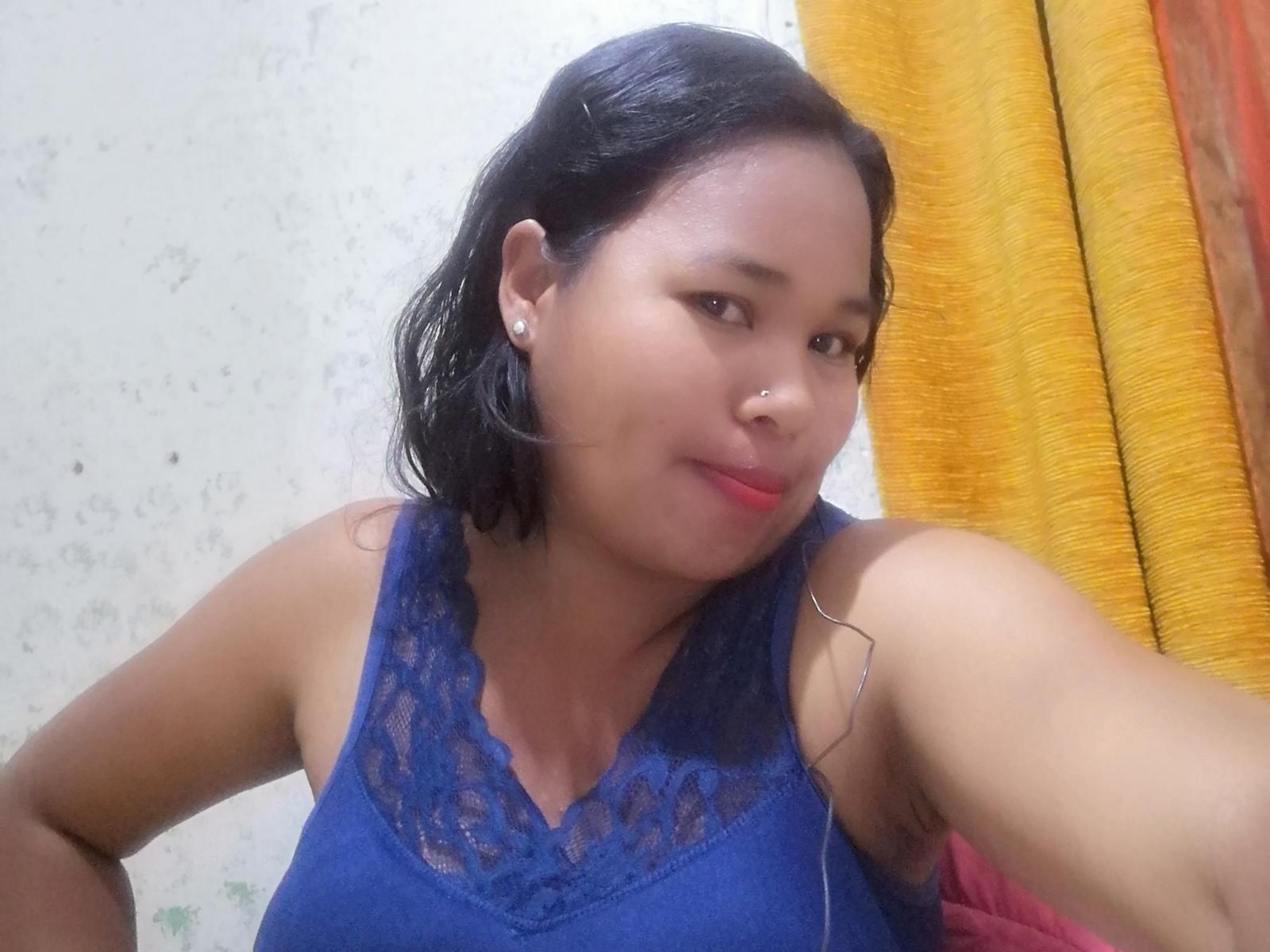 MoiraHot with medium brown skin, black shoulder-length hair, wearing a blue lace sleeveless top, small nose ring, pearl earrings, smiling slightly, yellow and orange curtain in background.