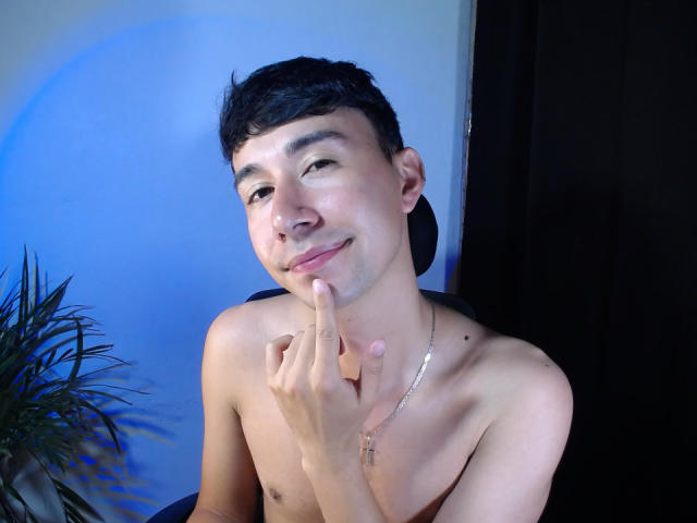 OliverHot69 is shirtless, with short black hair, a silver cross necklace, and a finger to his lips, smiling. His skin is light, and he has a cute, playful look.