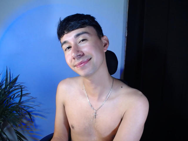 Shirtless OliverHot69 with short black hair, light skin, small cross necklace, slight smile, blue background, black curtain, plant on left. Smug, relaxed, upper body, casual.
