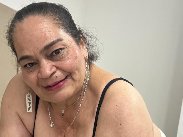 QueenMatturex, an older woman with dark hair in a ponytail, wearing a black spaghetti strap top, silver jewelry, and pink lipstick, smiling at the camera.