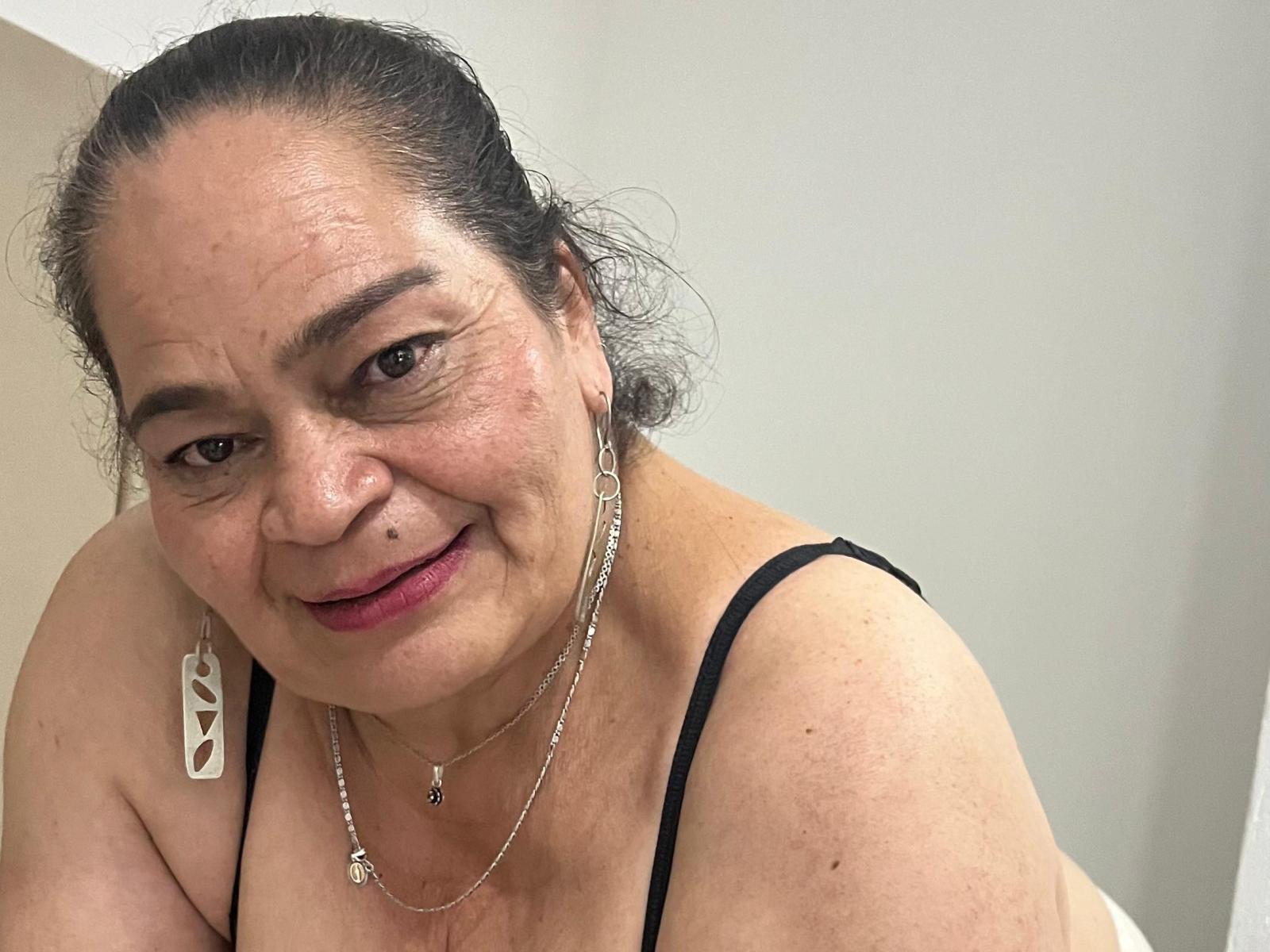 QueenMatturex, an older woman with dark hair in a ponytail, wearing a black spaghetti strap top, silver jewelry, and pink lipstick, smiling at the camera.