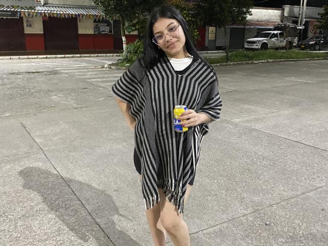Ebba with black hair, glasses, and a smile, wearing a black and white striped dress, holding a yellow and blue drink can, standing on a concrete surface.